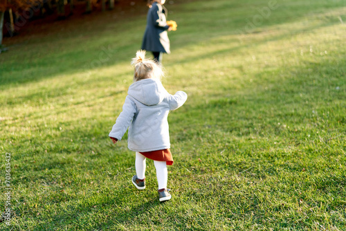 Little girl walks along a green lawn to her mother with a bouquet of yellow leaves. Back view