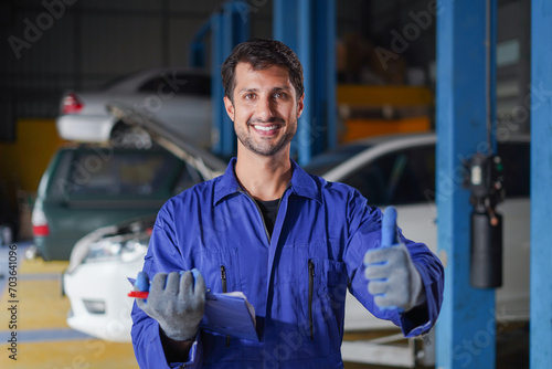 happy latin auto mechanic man checking car with clipboard gesturing show thumbs up and looking at camera in garage cars service . hispanic technician repairing vehicle at garage .