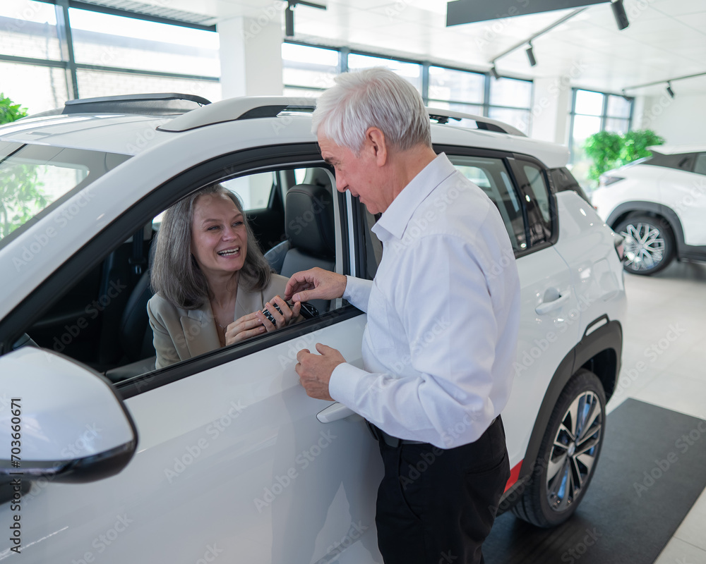 Mature Caucasian woman sits in a new car, an elderly man gives her the keys. Mature Caucasian woman sits in a new car, an elderly man gives her the keys.