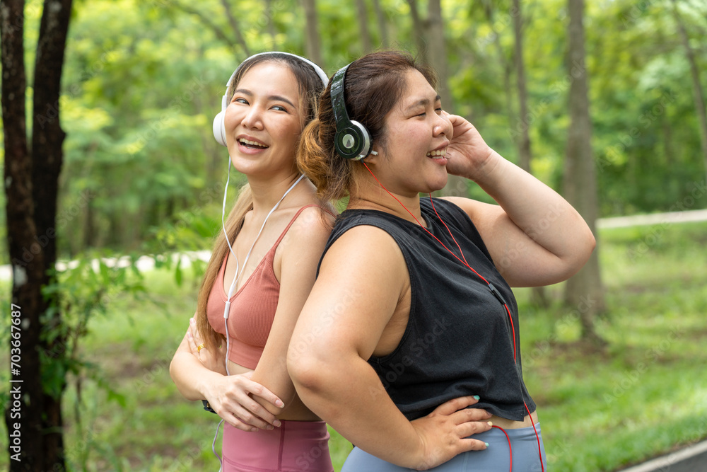Two happy Asian female friends, one over size and one lean, standing with their back together listening to music from their headphones at a local park Two happy Asian female friends, one over size and one lean, standing with their back together listening to music from their headphones at a local park