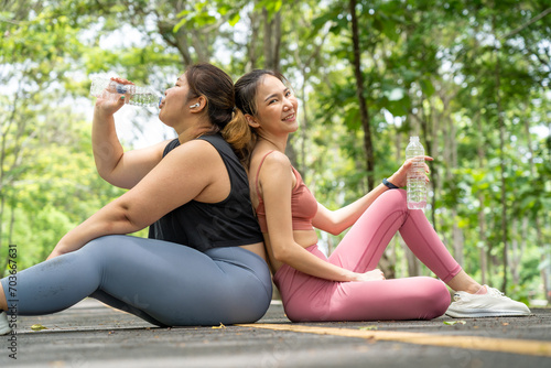 Two Asian women leaning their back together while sitting drinking water from plastic water bottle after their morning run at a local park