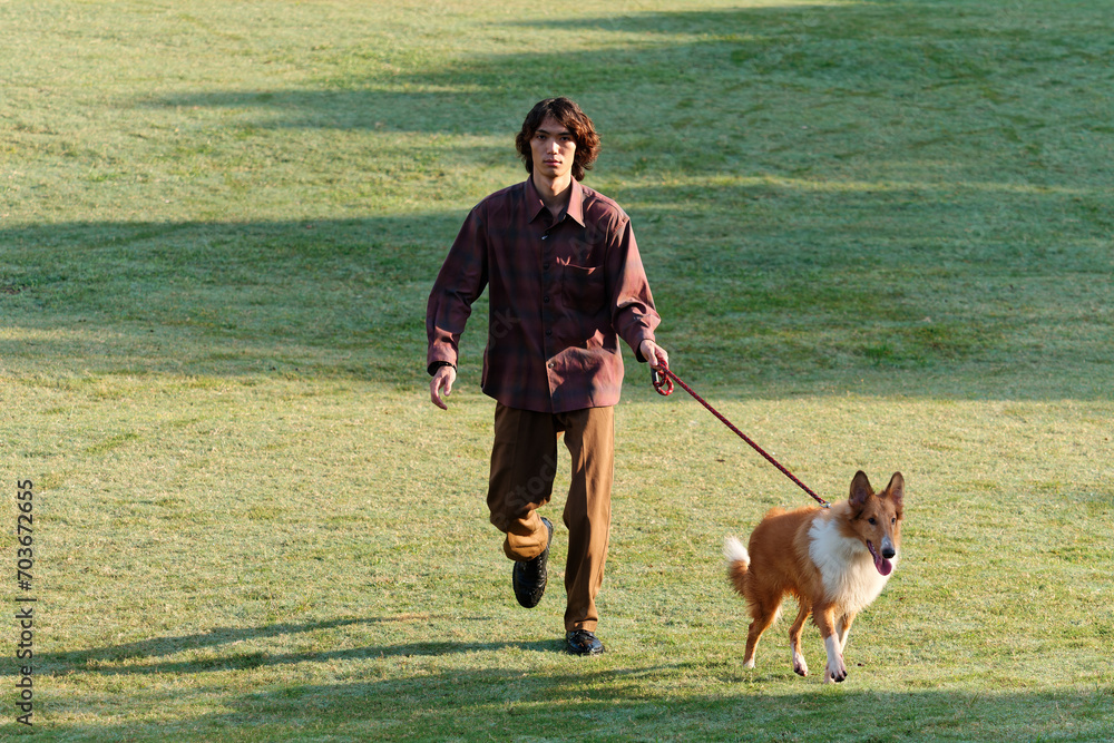 Portrait of handsome Chinese young man with curly hair running with his rough collie dog on green grass field in sunny day, male fashion, cool Asian young man lifestyle, harmony man and pet.