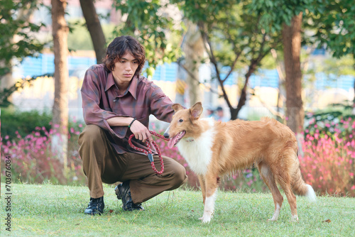 Portrait of handsome Chinese young man with curly hair squat with his rough collie dog on green grass field in sunny day, male fashion, cool Asian young man lifestyle, harmony man and pet. © atiger