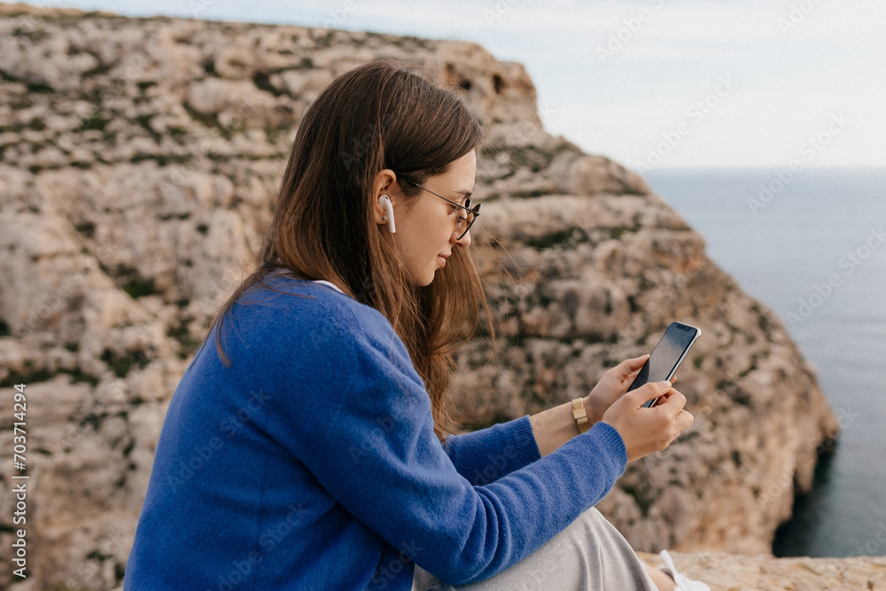 Profile photo of lovely cute girl on blue shirt wearing glasses and headphones using phone against mountains and ocean 