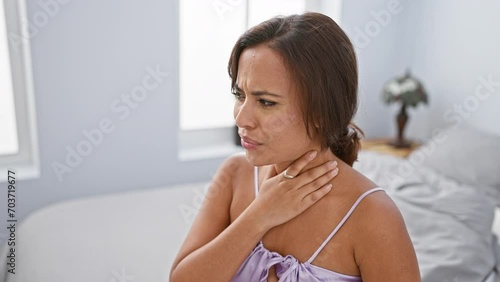 Young, beautiful hispanic woman in pyjamas sitting on her bedroom bed, in visible pain as she suffocates, strangled by unseen health problem. photo