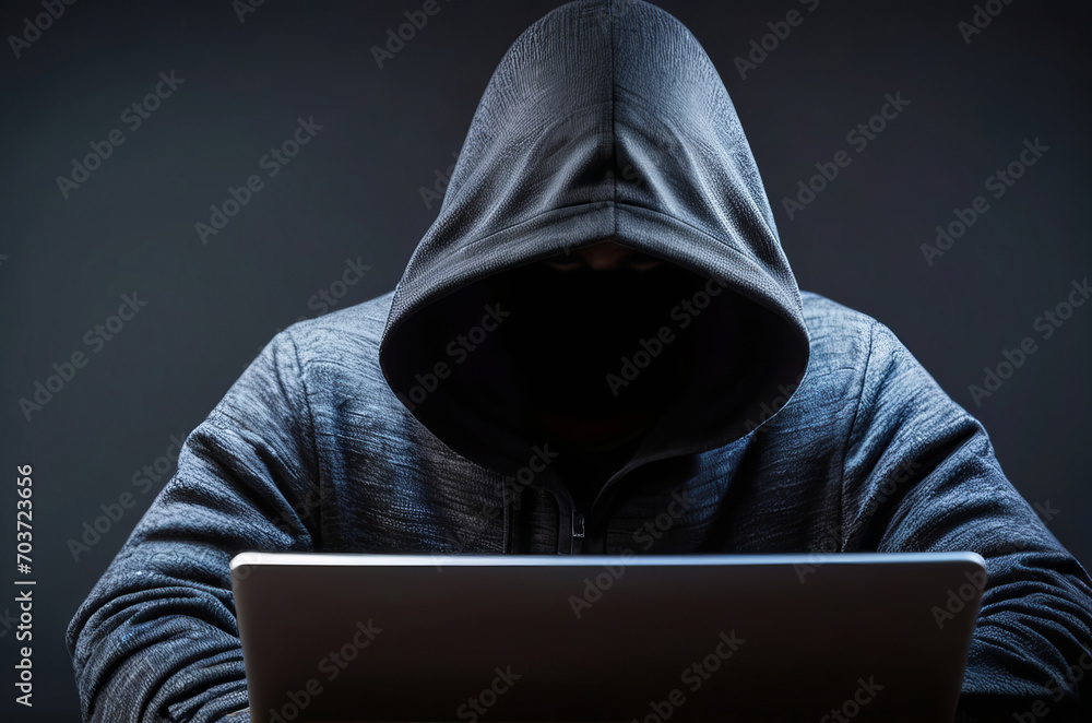 Cybersecurity Enigma: Hooded Figure at Laptop with Face Covered in the Shadows, Black Background. Cybersecurity Enigma: Hooded Figure at Laptop with Face Covered in the Shadows, Black Background.