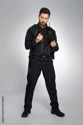 Collected caucasian police officer reloading weapon, getting ready to shoot. Front view of man in cop uniform recharging handgun, holding magazine, on gray studio background. Work, weapon concept.