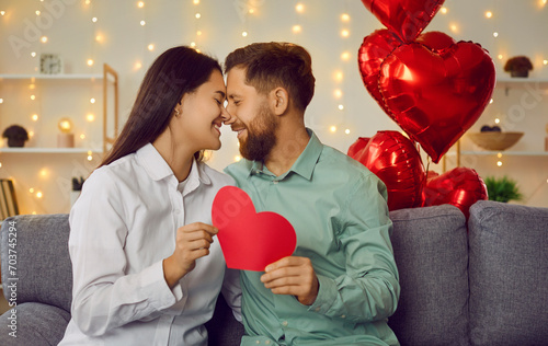 Happy couple holding red paper heart looking at each other. Romantic man and woman celebrating St. Valentine day sitting on background of red heart shaped balloons and glowing lights