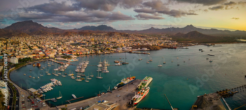 Panoramic aerial view of Mindelo city at sunset, with the marina and boats in the foreground, surrounded by the vibrant cityscape and mountains in the background under a warm, glowing sky