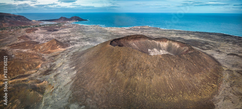 Aerial view of a coastal volcanic landscape (Viana volcano, Sao Vicente) with a distinct crater in the foreground, sparse vegetation, and a small village by the sea, under a cloudy sky