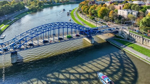 Blue tied-arc Pilsudski bridge over Vistula river in Krakow, Poland. A tram and cars passing the bridge. Tourist ship sailing under it. Flood walls and boulevards on the riverbanks. Aerial 4K video photo