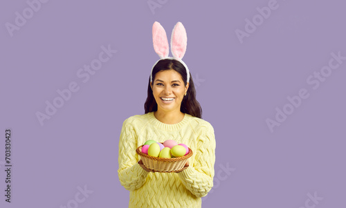 Studio portrait of pretty bunny girl. Happy cheerful woman in yellow sweater and cute rabbit ears standing isolated on purple background, looking at camera, smiling and showing bowl of Easter eggs