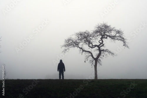 A minimal edit of a eerie spooky figure, back to camera, alone. Standing next to a tree on a creepy foggy winters day.