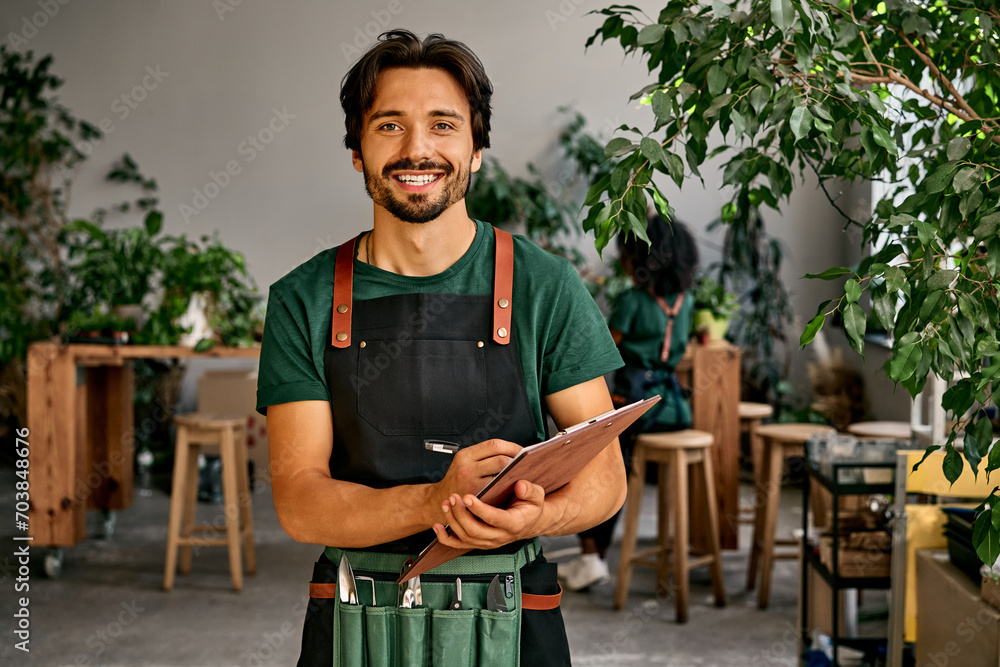 Handsome confident sincere smiling male gardener florist in green t-shirt black apron and bag of garden tools on belt holding wooden clipboard and pen looking at camera. Work in a plant shop. - obrazy, fototapety, plakaty 
