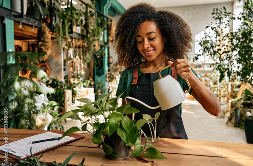 Love for plants. African american female gardener in green apron standing at flower store and using watering can. Positive young woman with curly hair taking care of greenery in pots. photo