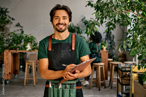 Handsome confident sincere smiling male gardener florist in green t-shirt black apron and bag of garden tools on belt holding wooden clipboard and pen looking at camera. Work in a plant shop. photo