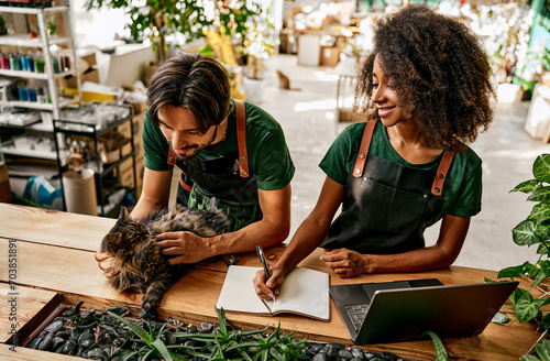 Atmospheric floral boutique. Attractive woman in apron writing in copybook and using laptop while man caressing dark cat at flower shop. Multiethnic family running own small business with love. photo