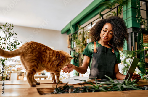 Beautiful confident successful business lady in green t-shirt and black apron petting red cat while working at computer in plant shop. A pet at work with the owner.