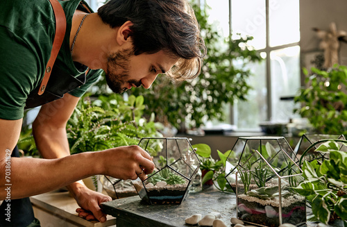 A handsome man makes a composition in a glass florarium, holds stones with tweezers. Work in a store of plants, decor, florariums.