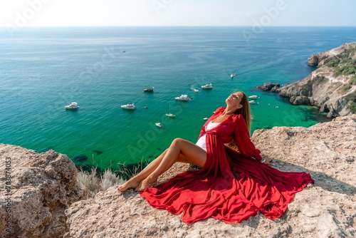 Woman red dress sea. Happy woman in a red dress and white bikini sitting on a rocky outcrop, gazing out at the sea with boats and yachts in the background.