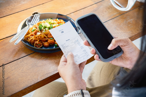 Closeup woman is holding a food receipt and smart phone in the restaurant, scan, or online payment, calculate the food price, focus at the receipt photo