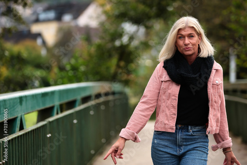 Woman blonde in her fifties with a pink denim jacket, blue jeans with a ripped black sweater and a black scarf, portrait on a bridge.