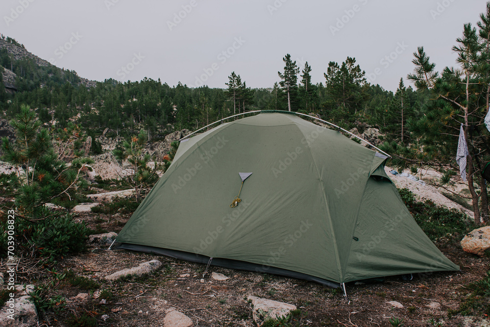 Green tourist tent set up in a forest surrounded by evergreen conifers and rocks in a mountainous area.