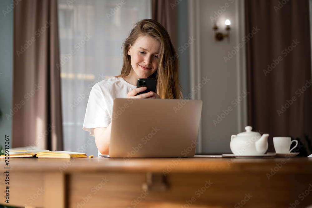 Smiling young woman using smartphone behind laptop, with a teapot and cup, in a warm home setting. Smiling young woman using smartphone behind laptop, with a teapot and cup, in a warm home setting.