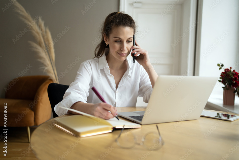 Professional woman multitasking with a phone call and note-taking, organized at her laptop. Professional woman multitasking with a phone call and note-taking, organized at her laptop.
