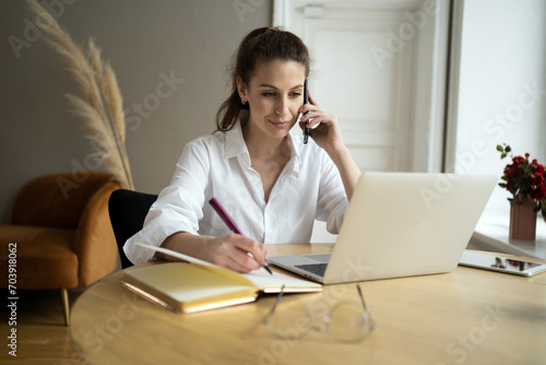 Professional woman multitasking with a phone call and note-taking, organized at her laptop.