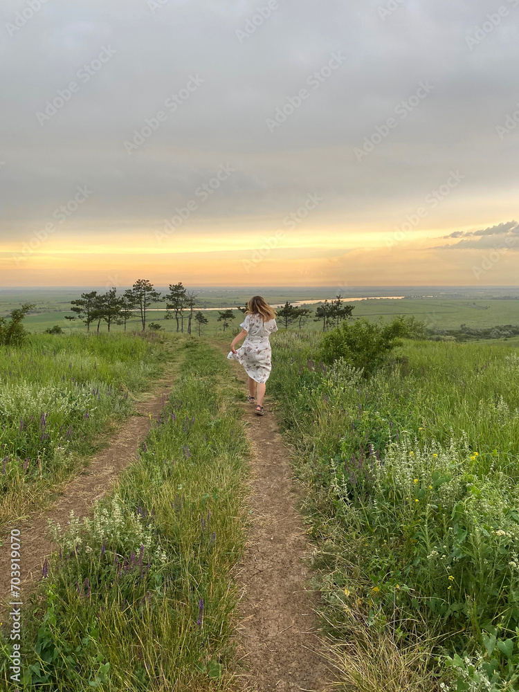 Back view of woman in romantic dress running through field of green grass at sunset on summertime
