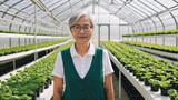 Smiling senior asian female with gray hair and eyeglasses looking at camera while standing with green vest and white shirt in modern white greenhouse with organic hydroponic farm and green plants