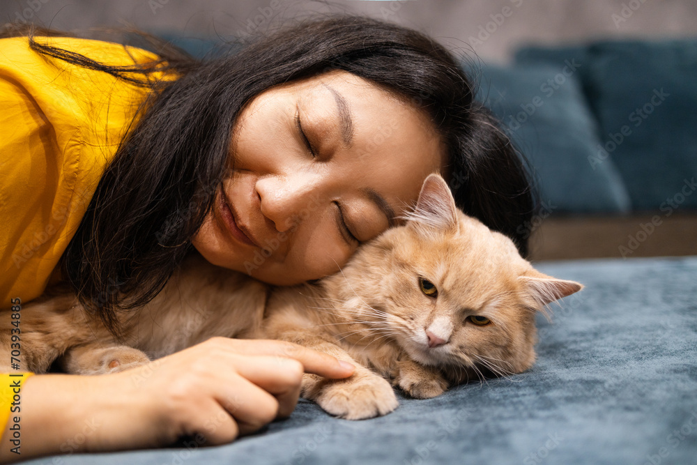 Close up beautiful Asian woman lie near her orange cat also scratch the chin of cat to make it relax and happy on bed.