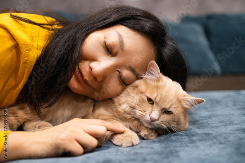 Close up beautiful Asian woman lie near her orange cat also scratch the chin of cat to make it relax and happy on bed.