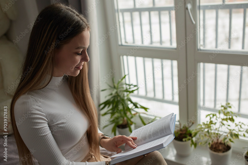 Young woman sitting on modern sofa in front of window relaxing in her living room reading book Young woman sitting on modern sofa in front of window relaxing in her living room reading book