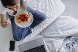 Top view of Asian Thai woman eating homemade pasta and sitting on sofa bed in room apartment while watching series movie by laptop, using notebook working alone in day time.