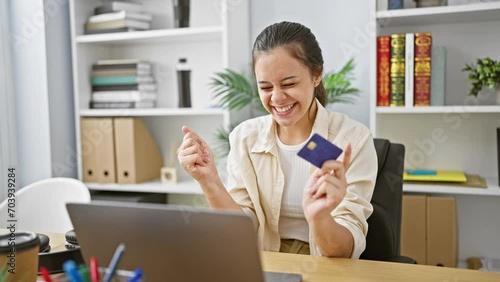 Victorious young hispanic woman celebrating financial success in office, beautiful business worker exults in big win while working online, laptop open, credit card at hand! photo