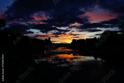 Bild auf Leinwand In this mesmerizing landscape, a tranquil river reflects the warm hues of a setting sun, harmonizing with the silhouette of an bridge against the vibrant sky