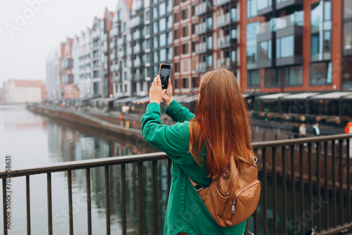 Back view of young European tourist makes photo or video on smartphone at city of Gdansk, Poland. Hipster girl with backpack hold on smart phone gadget outdoors © mdyn
