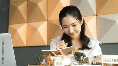 Young beautiful asian architect taking a note while inspect architectural model at modern office. Professional project manager checking house model during writing on blueprint at office. Manipulator.