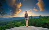 Sportive woman standing alone on hillside trail. Female hiker enjoying view of evening nature from rocky cliff on wilderness path. Active lifestyle concept