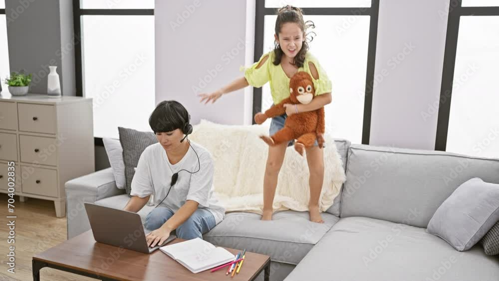 Mother working from home, distracted by daughter joyfully jumping on the sofa, disturbing peaceful indoor family moment Mother working from home, distracted by daughter joyfully jumping on the sofa, disturbing peaceful indoor family moment