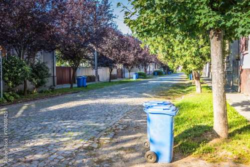 Fototapeta Naklejka Na Ścianę i Meble -  Blue garbage cans on the street in a small town near every house. Centralized garbage collection in a small cozy European city. Garbage collection on a certain day of the week.