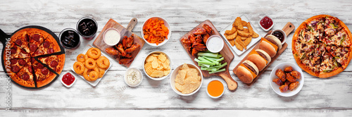 Junk food table scene. Pizza, hamburgers, chicken wings and salty snacks. Top view over a white wood banner background. photo