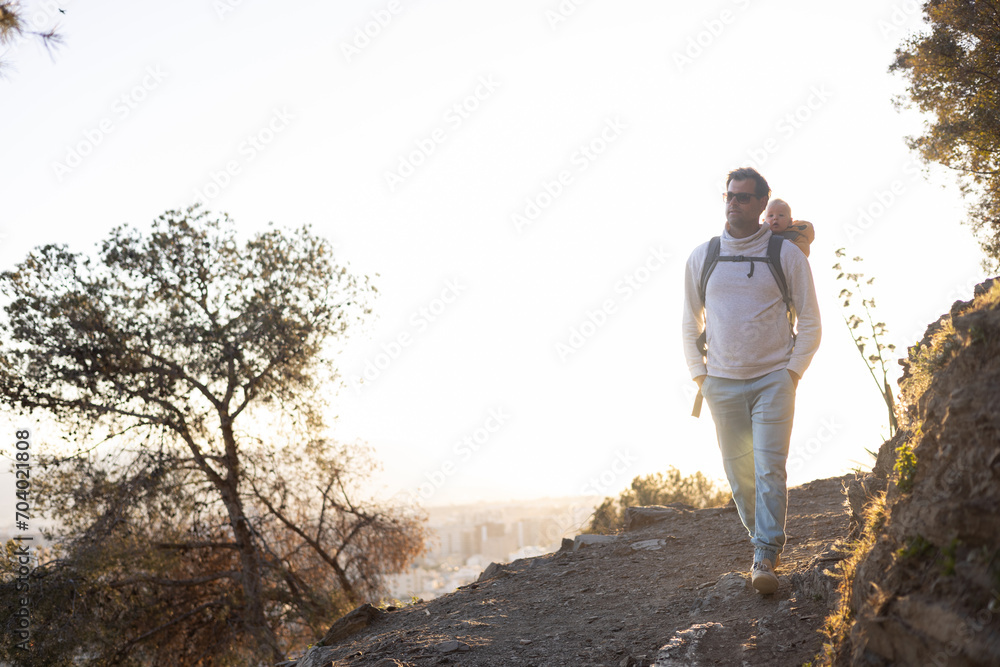 Young father carrying his infant baby boy son in backpack while tracking around Malaga, Spain in sunset. Family travel and vacation concept.