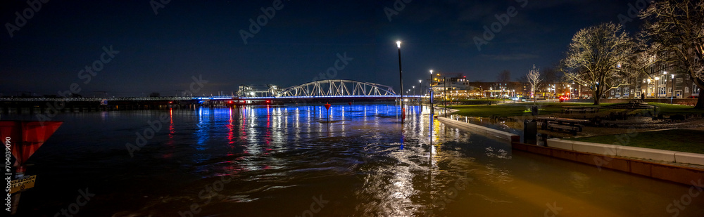 Wide panorama of colourful decoration on steel draw bridge in the background with high water levels of river IJssel flooded countenance boulevard of tower town Zutphen at nighttime with street lights