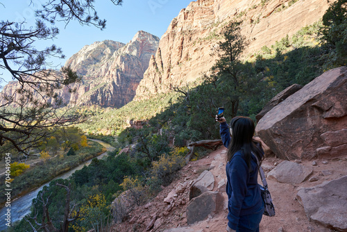 A tourist explores a trail in zion national park with her phone camera. She snaps a picture of the virgin river and nearby mountain peaks.