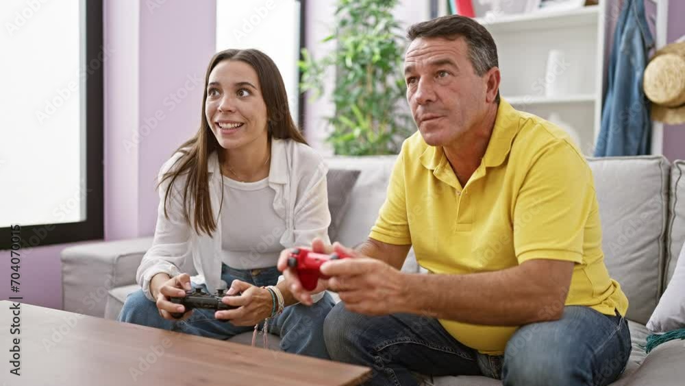 Hispanic father and daughter enjoy bonding time playing a video game at home, sitting on the living room sofa, smiles of joy on their faces, as they confidently use their gamepad controls. Hispanic father and daughter enjoy bonding time playing a video game at home, sitting on the living room sofa, smiles of joy on their faces, as they confidently use their gamepad controls.