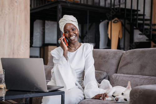Smiling African woman in a headscarf having a pleasant conversation on a mobile phone stroking dog at home. Girl in turban enjoying phone talk with friend. photo