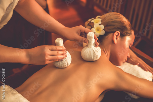 Hot herbal ball spa massage body treatment, masseur gently compresses herb bag on woman body. Tranquil and serenity of aromatherapy recreation in warm lighting of candles at spa salon. Quiescent photo